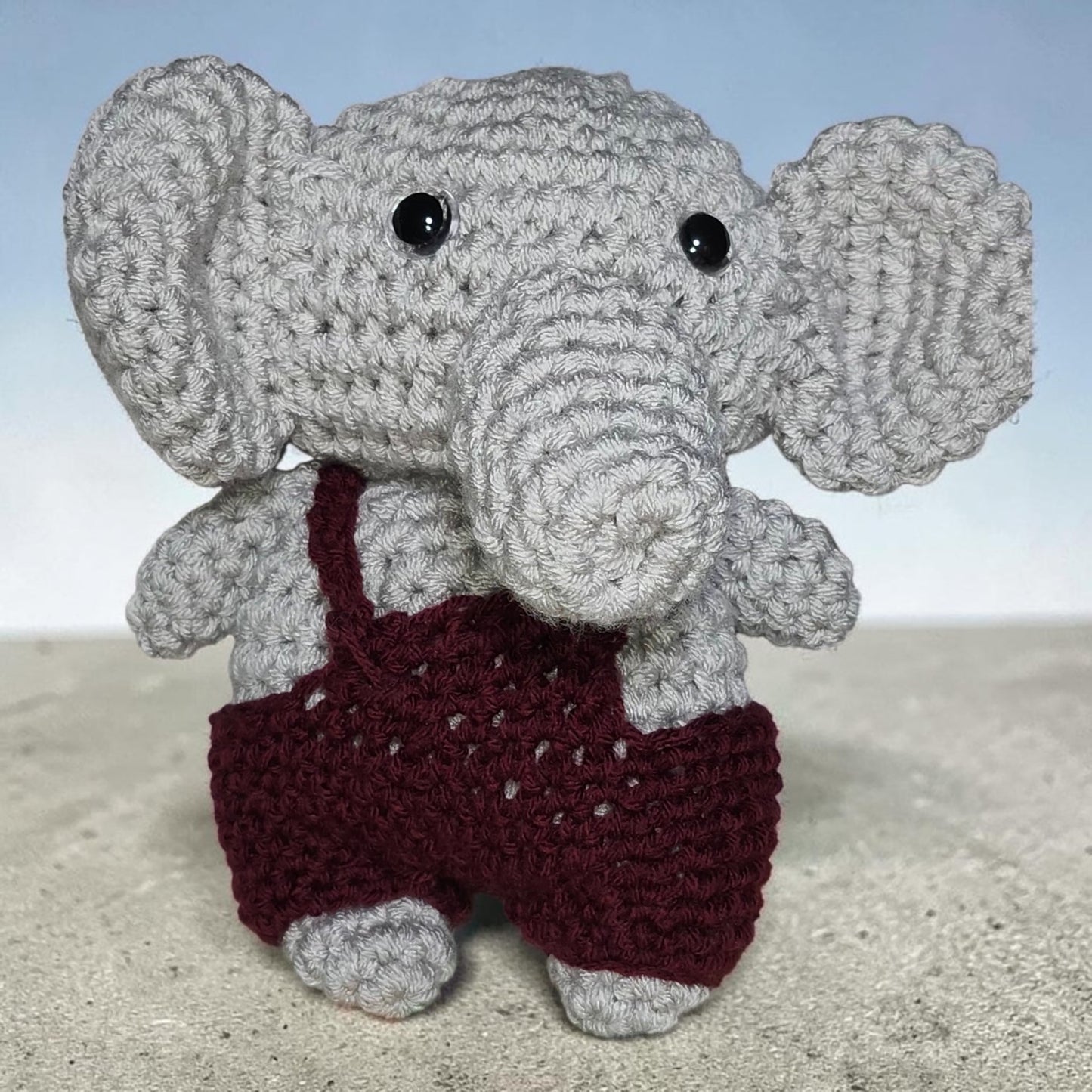 Handmade crochet elephant wearing overalls displayed against a neutral background.